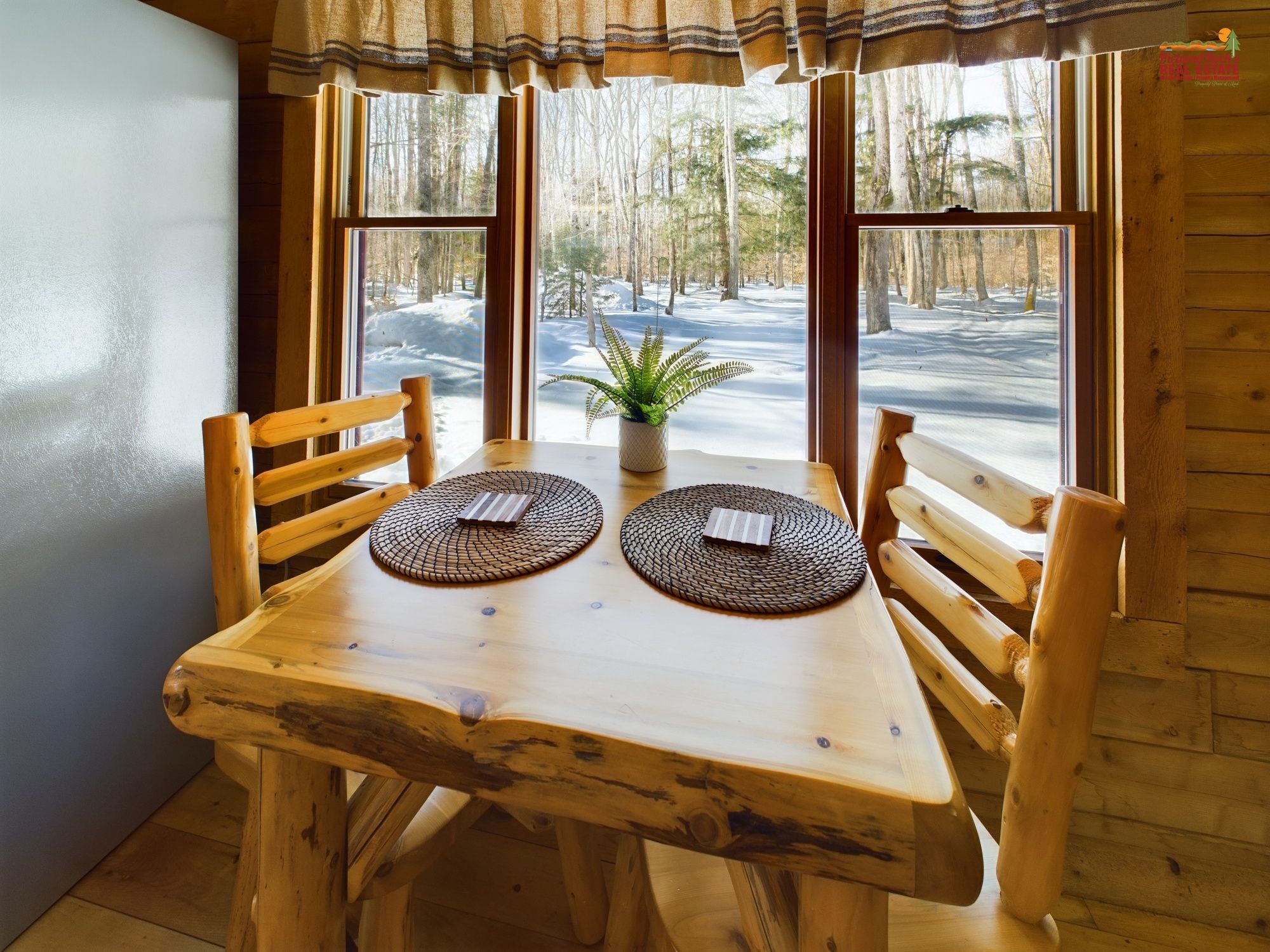 A wooden dining table with two placemats and a potted plant, set against a window with a snowy forest view.