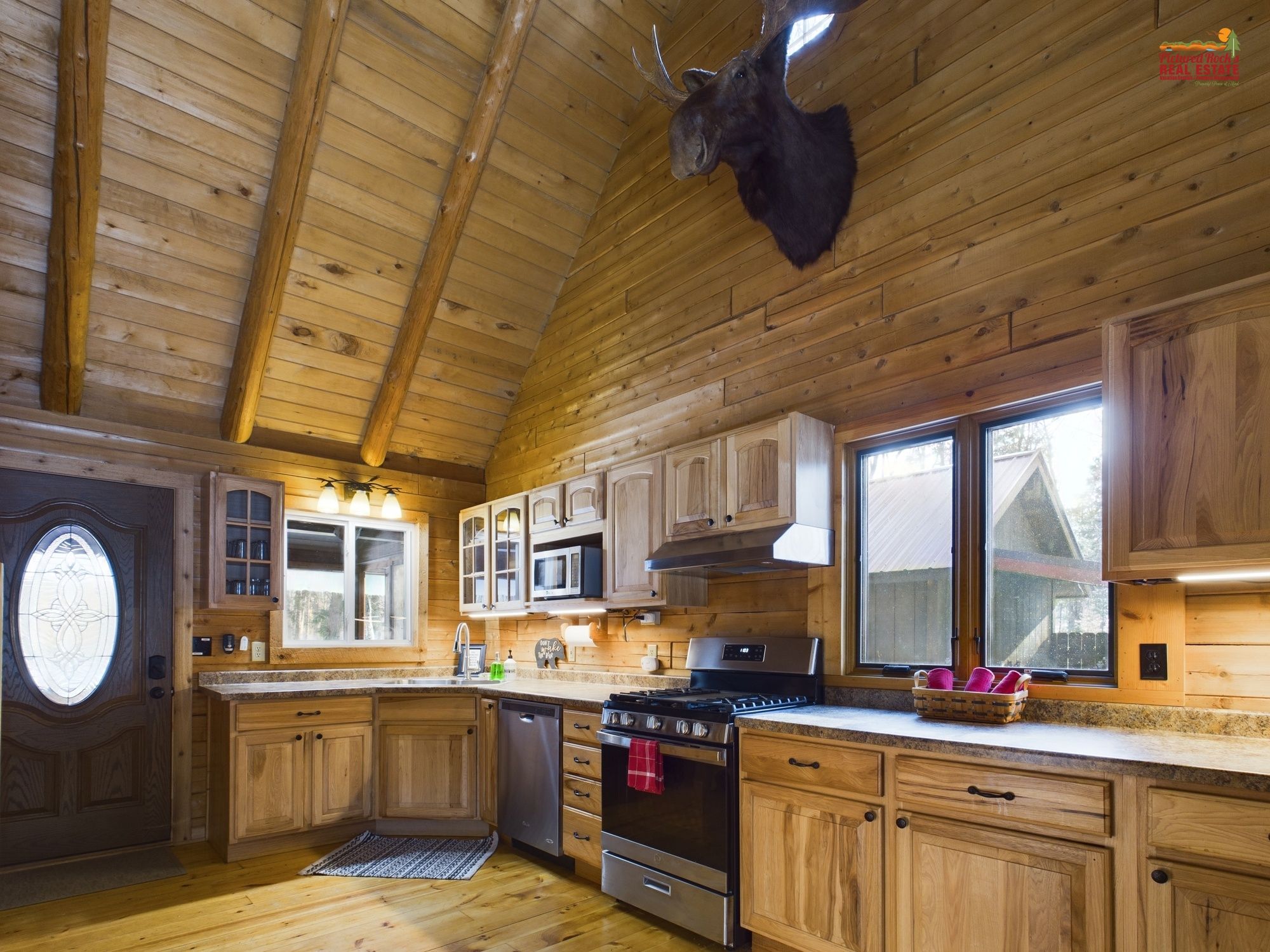 A wooden cabin interior with a kitchen featuring wooden cabinets, a stove, and a mounted moose head.