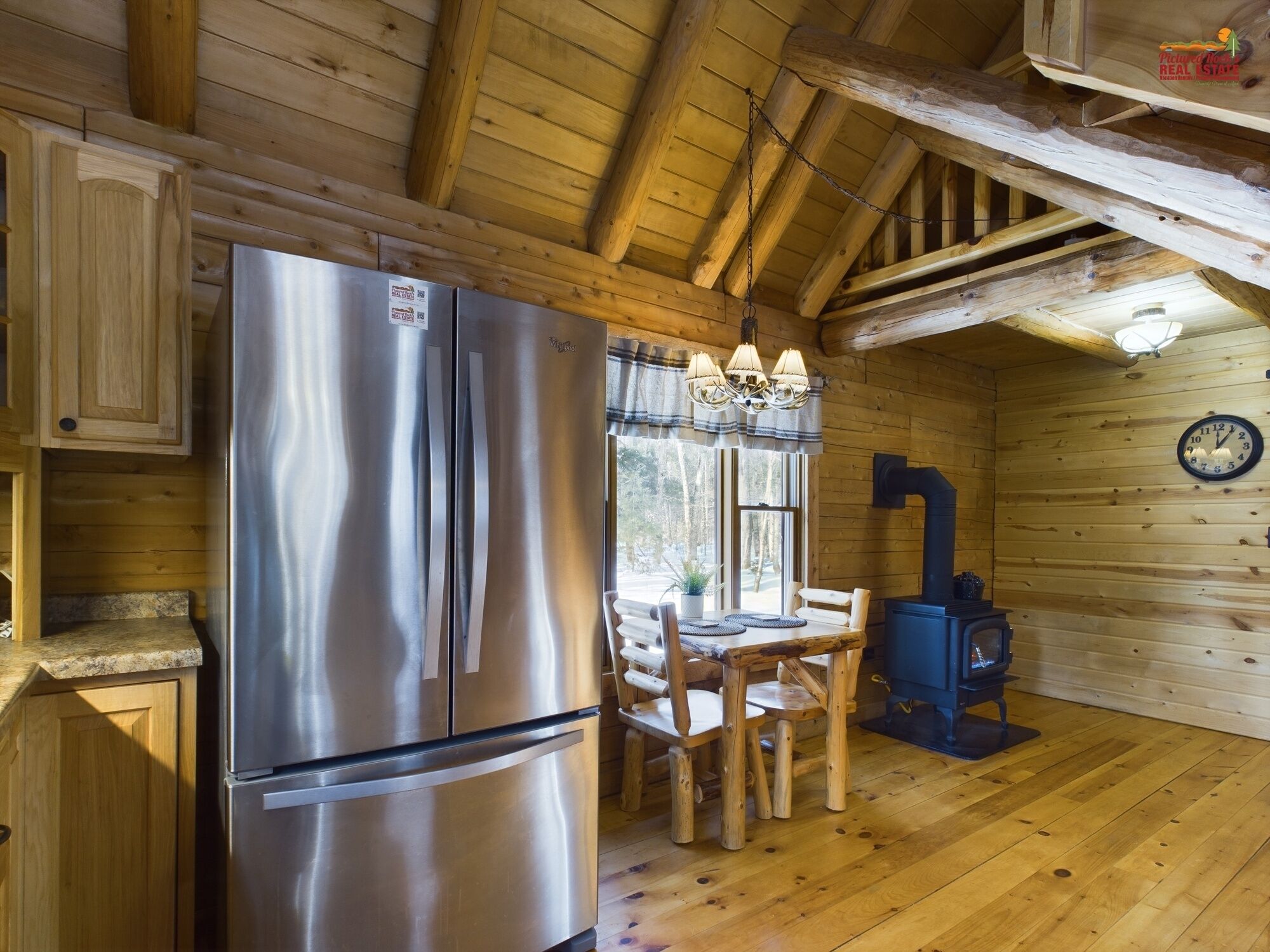 Interno di una baita in legno con frigorifero in acciaio inossidabile, tavolo da pranzo con sedie, lampadario e stufa a legna.