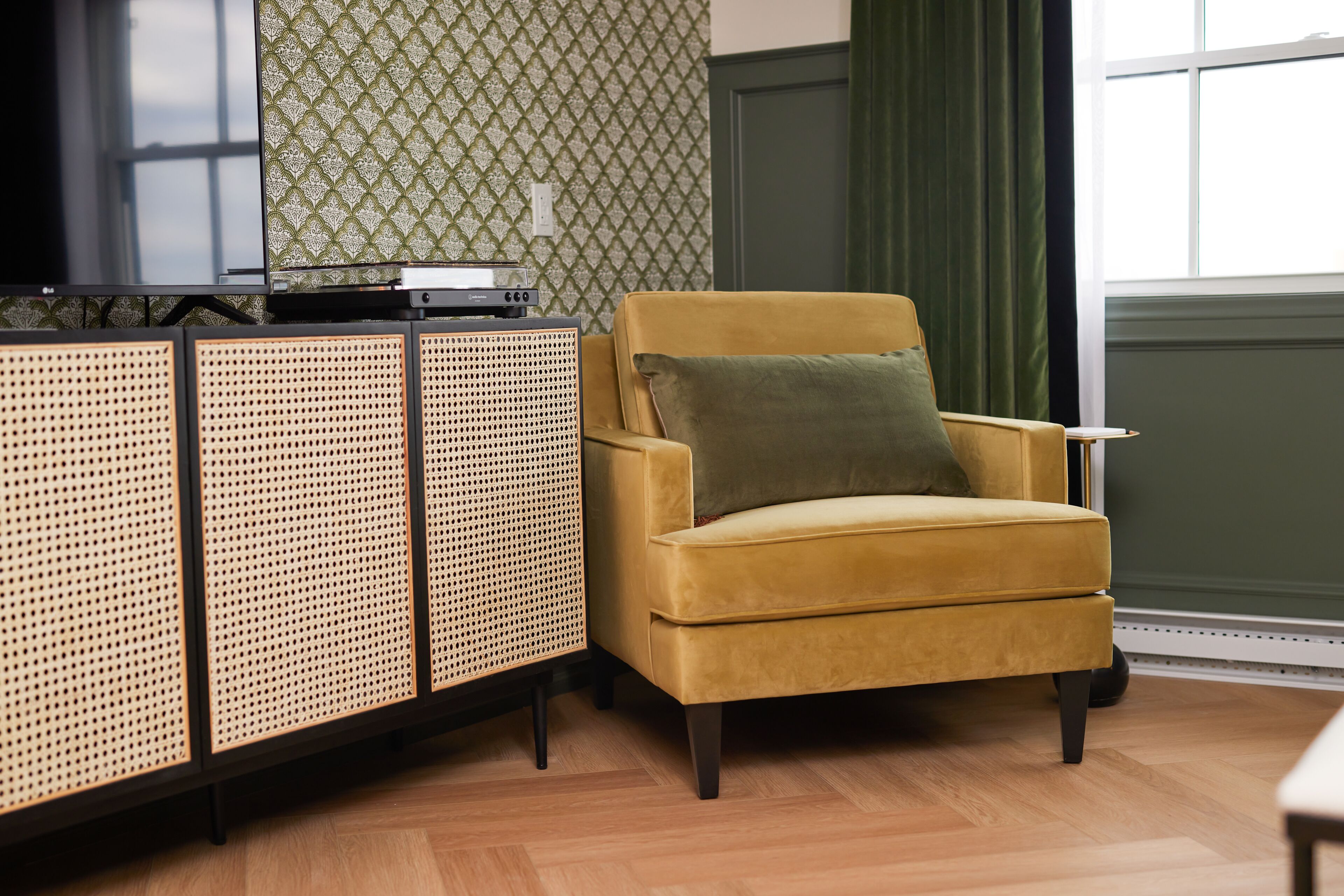 A room with a yellow armchair, a woven sideboard, and a patterned wallpaper.