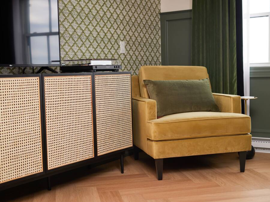 A room with a yellow armchair, a woven sideboard, and a patterned wallpaper.