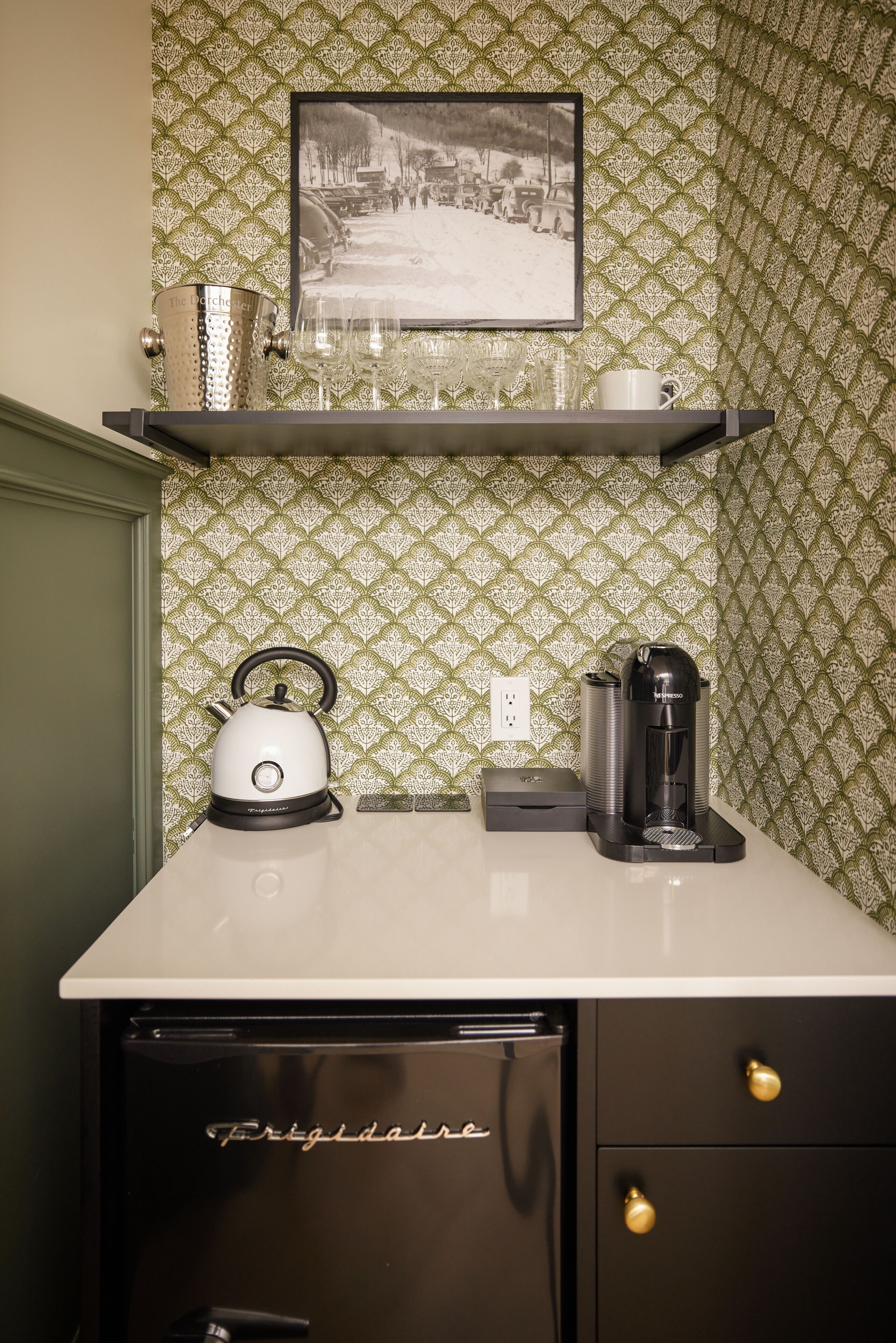 A kitchen with a Frigidaire refrigerator, a kettle, a coffee maker, and a shelf with glasses and a mug.