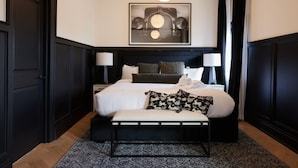 Un dormitorio moderno con una cama grande, dos mesitas de noche, un banco, un tapete estampado y una obra de arte enmarcada sobre la cama.