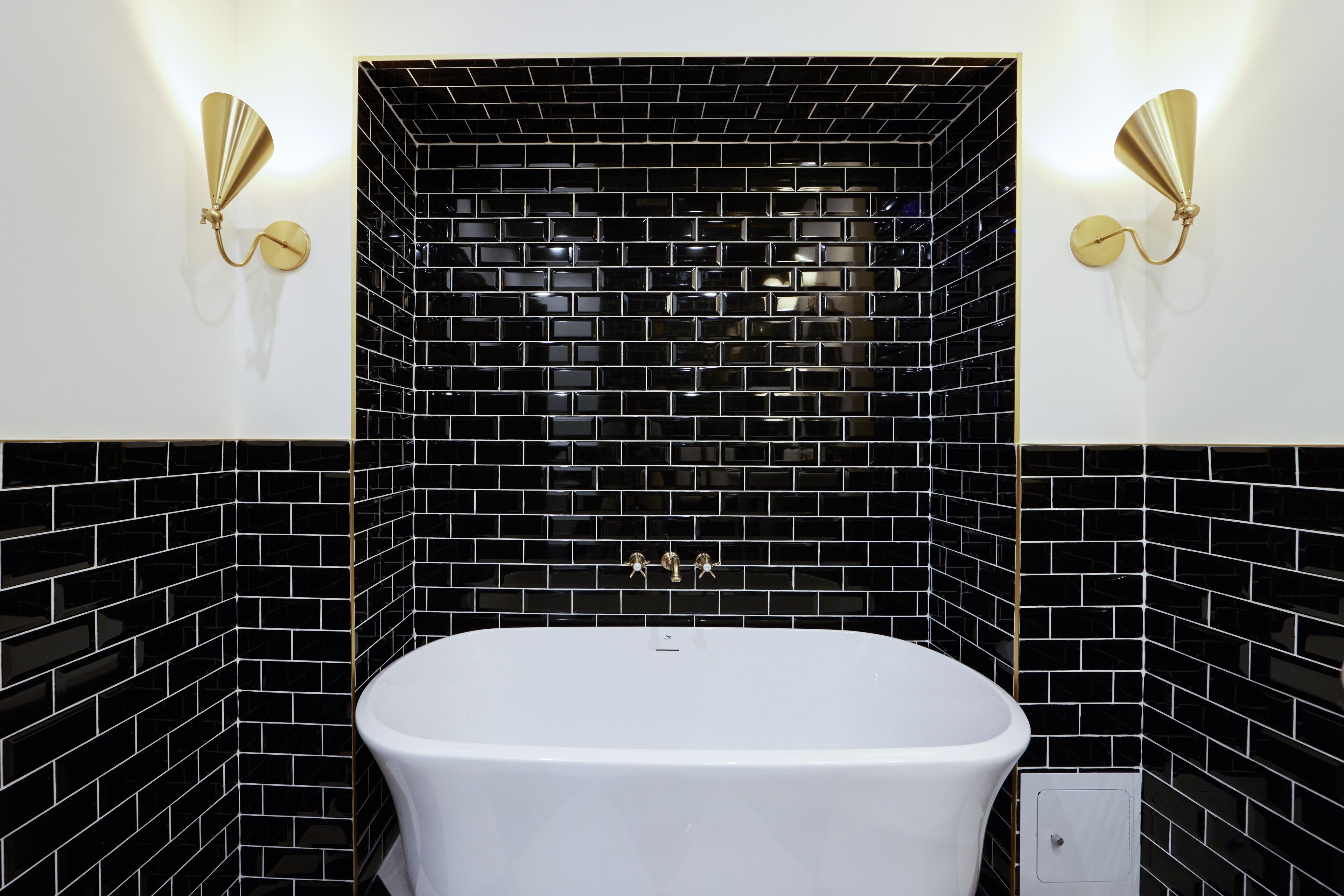 Un baño con una bañera blanca, paredes cubiertas de azulejos negros y dos luces empotradas.