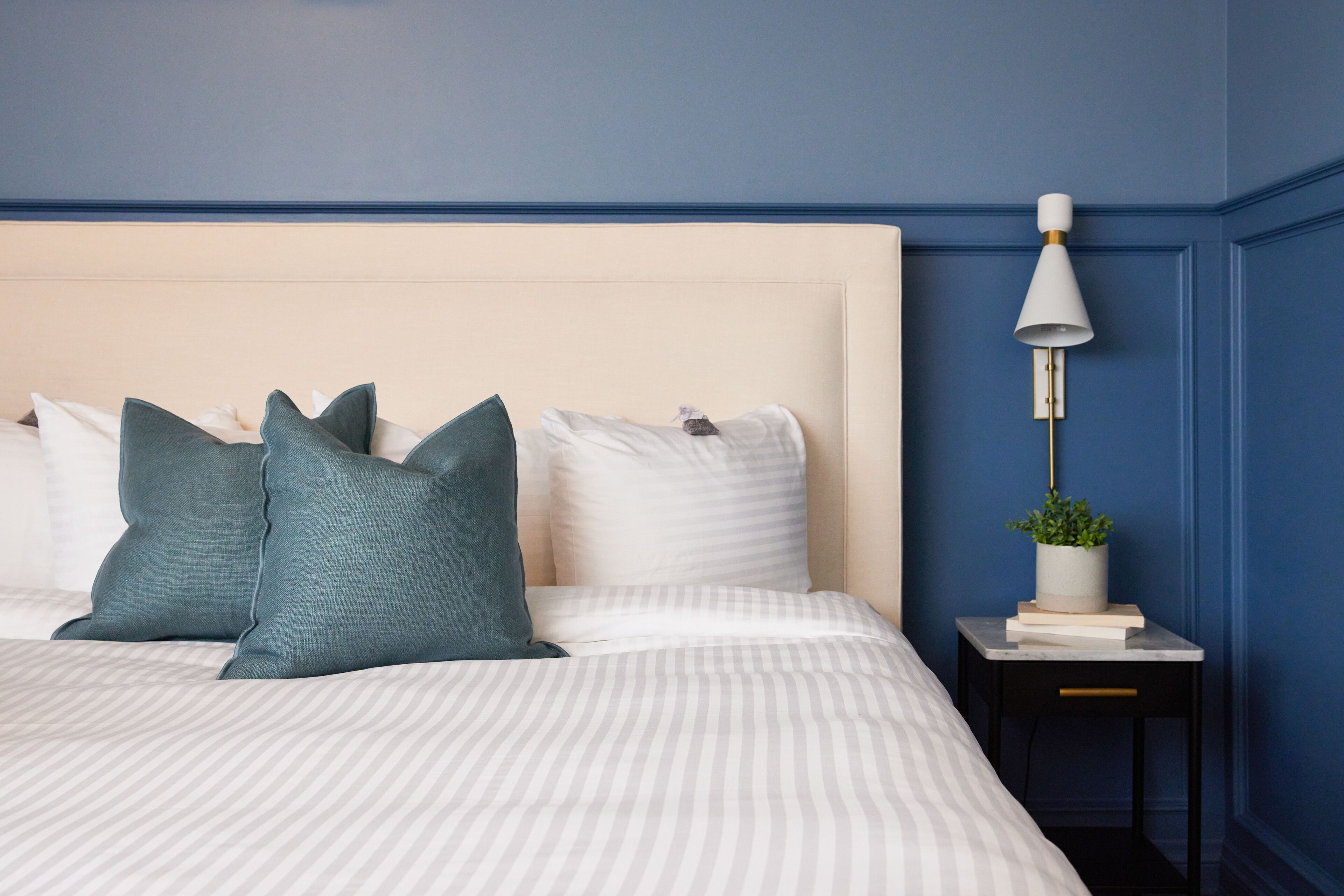 Una cama bien hecha con ropa de cama a rayas y almohadas color turquesa, una mesita de noche con una lámpara y una planta en maceta, y una pared azul.