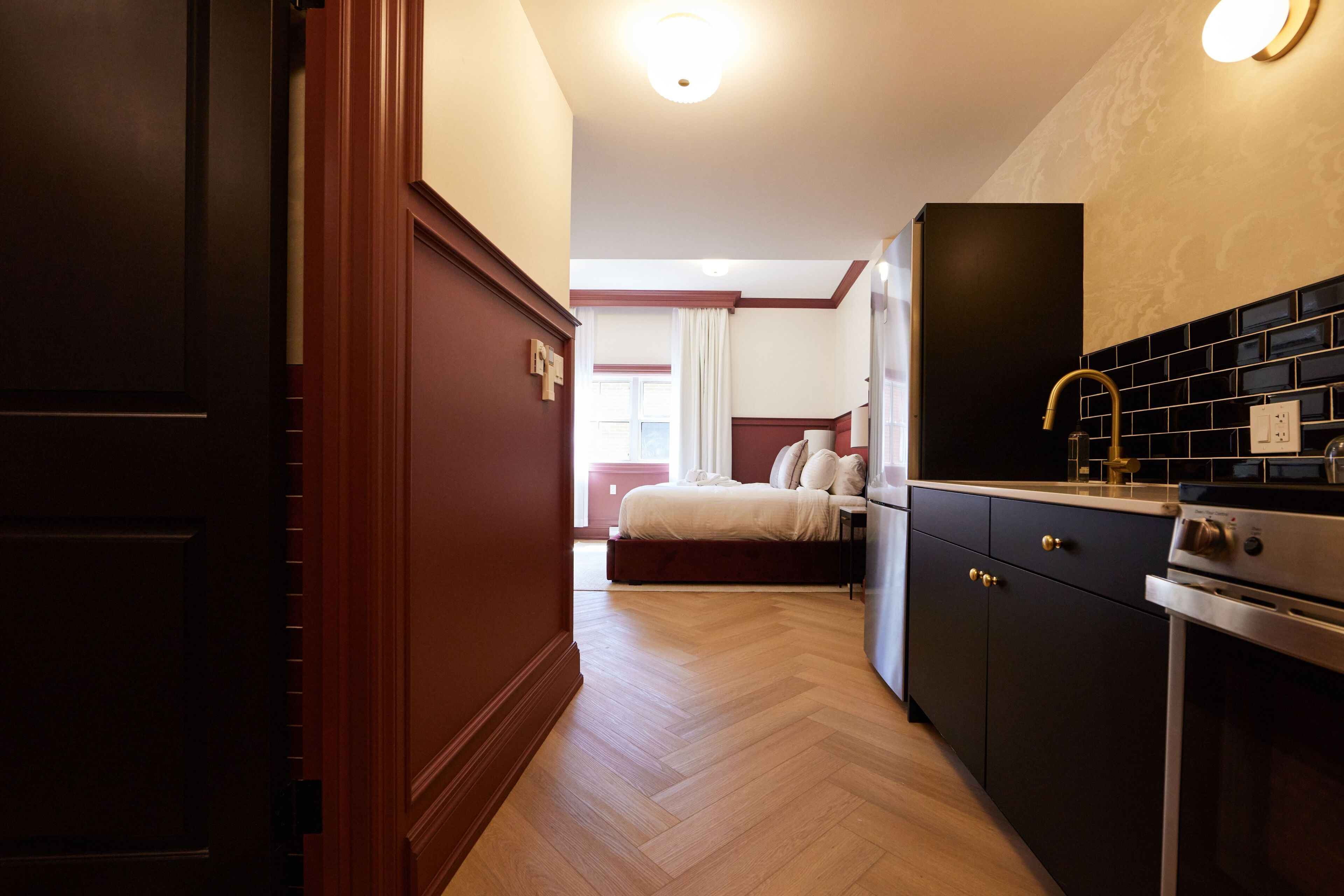 A hotel room with a wooden floor, a bed with white bedding, a dark wooden wardrobe, a kitchen area with a stove, and a window with curtains.