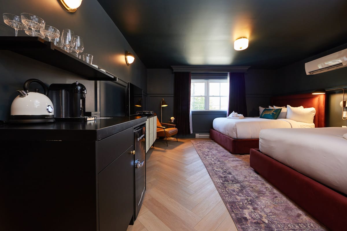 Ett hotellrum med ett litet kök, en säng med en röd sänggavel, ett fönster med gardiner och en mönstrad matta.