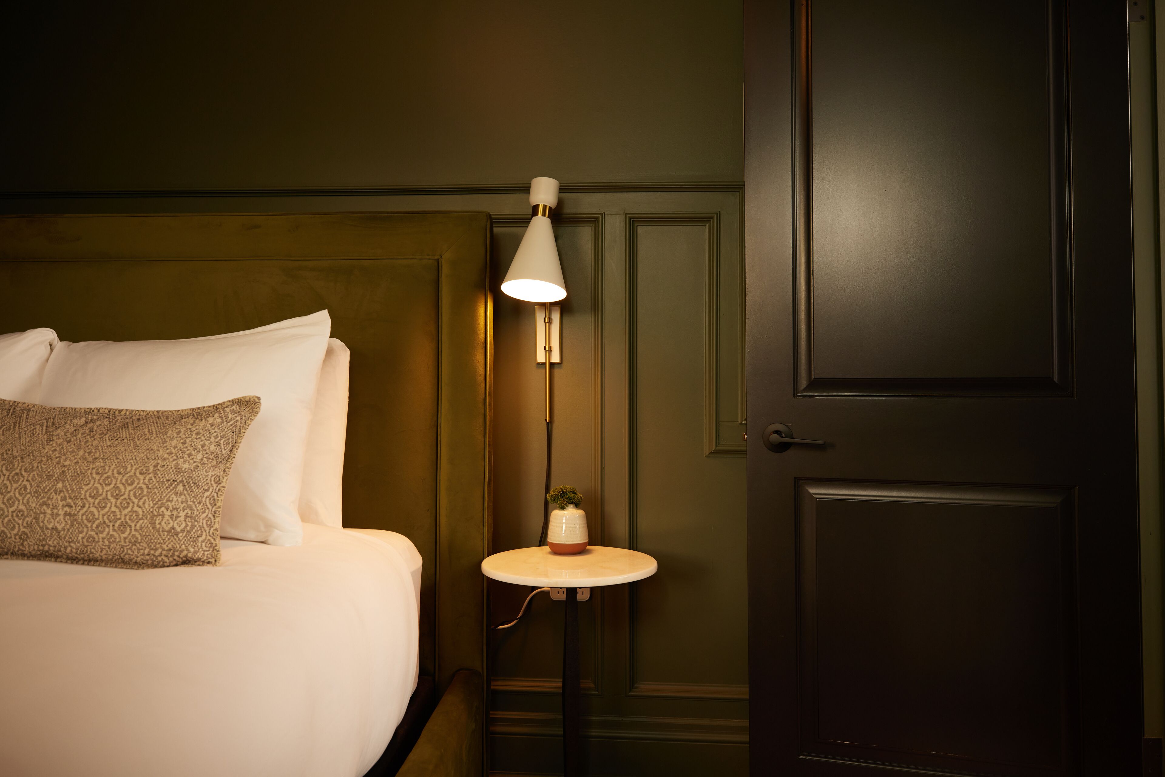 A neatly made bed with a pillow, a bedside table with a lamp, and a door in a room with dark walls.