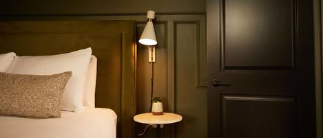 A neatly made bed with a pillow, a bedside table with a lamp, and a door in a room with dark walls.