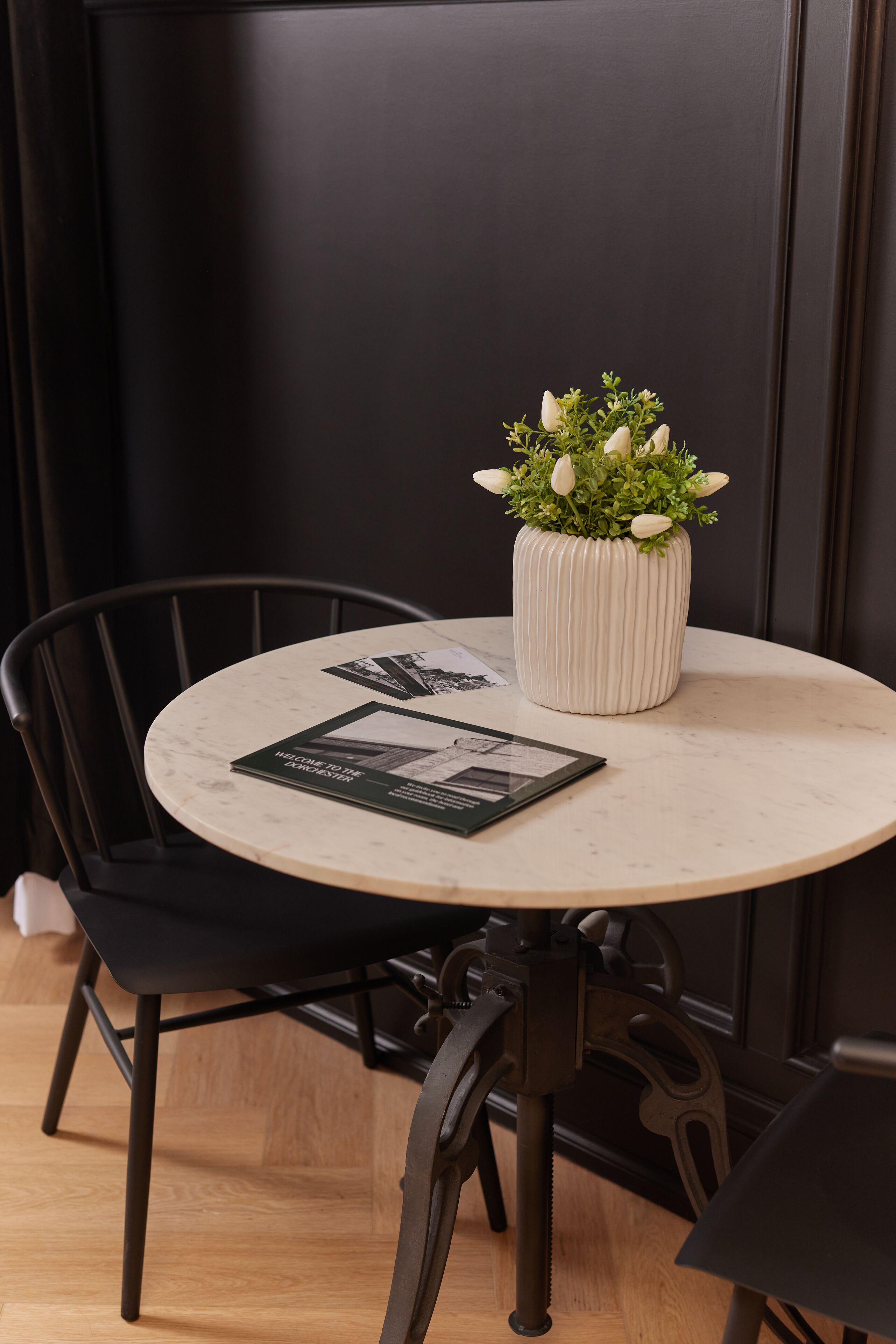 A round table with a white marble top, a white ribbed vase with green plants, and a black chair with metal legs.