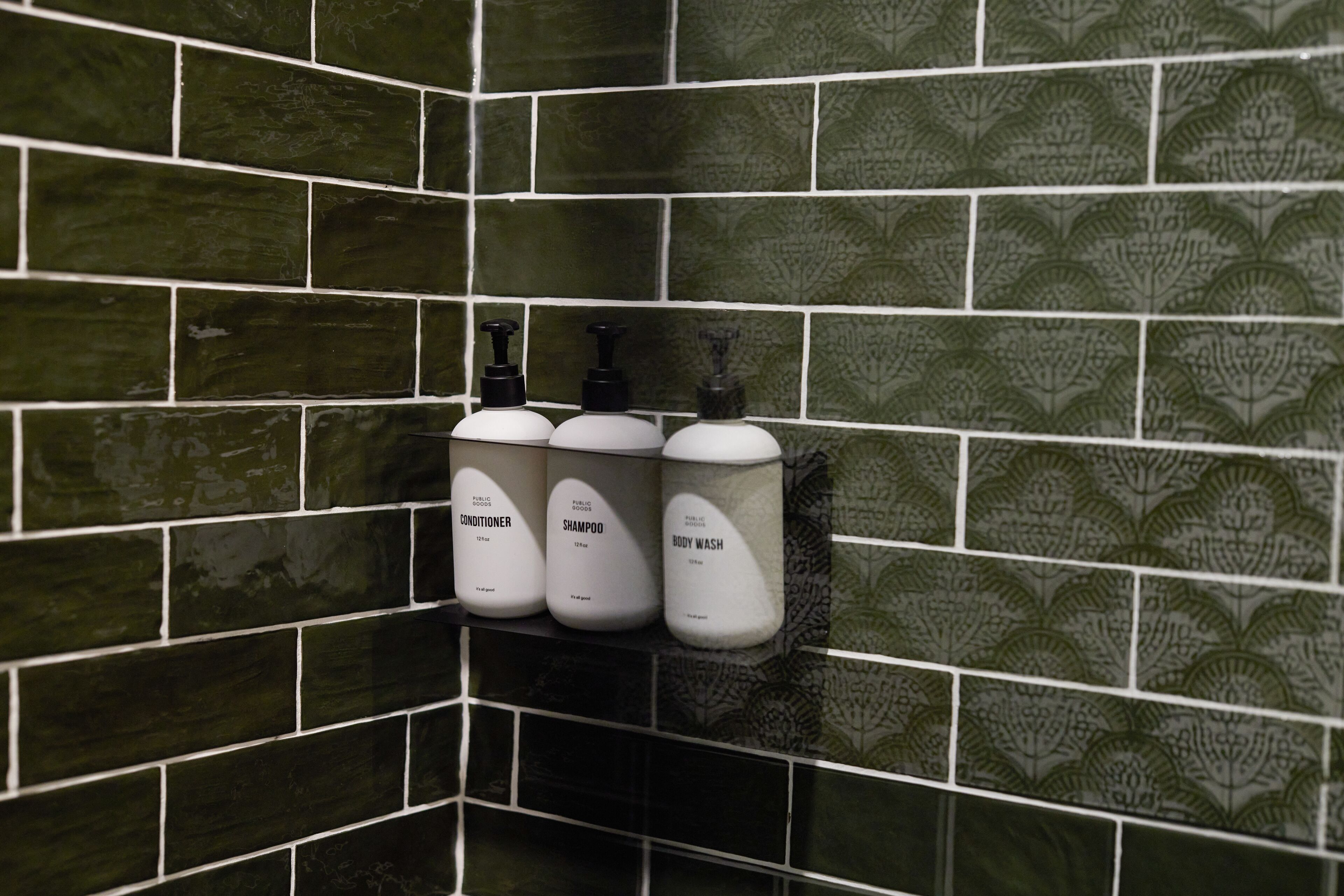 A bathroom with green patterned tiles and three white dispensers labeled conditioner, shampoo, and body wash.