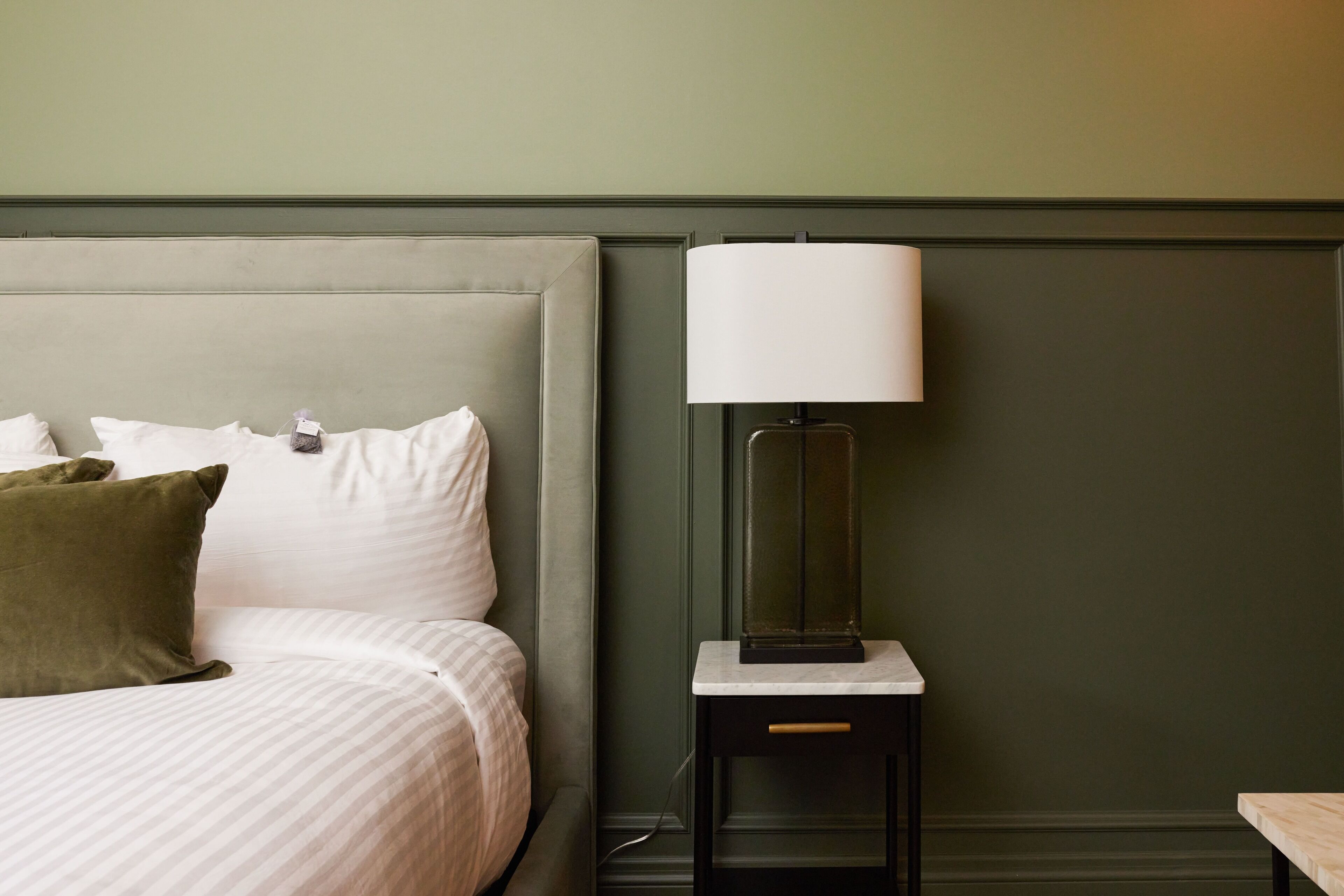 A neatly made bed with white linens and a green pillow, a bedside table with a lamp, and a green wall.