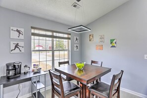 Un comedor con una mesa de madera, cuatro sillas, una cafetera y un cuenco de fruta sobre la mesa.