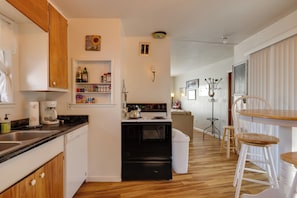 Efficient kitchen with oven, stove, and sink, adjacent to a dining area with a wooden table and two bar stools.