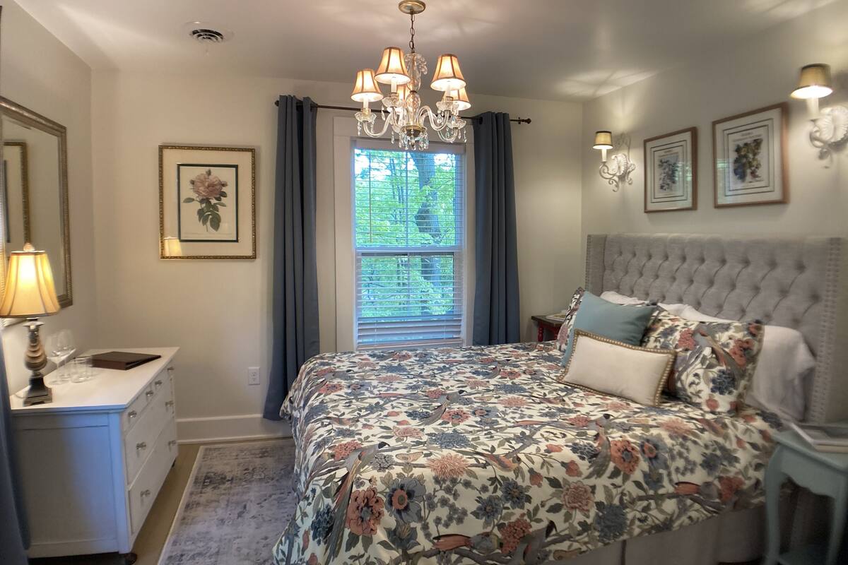 Une chambre à coucher comprenant un lit, une commode, une table de chevet, un lustre et une fenêtre avec des rideaux.