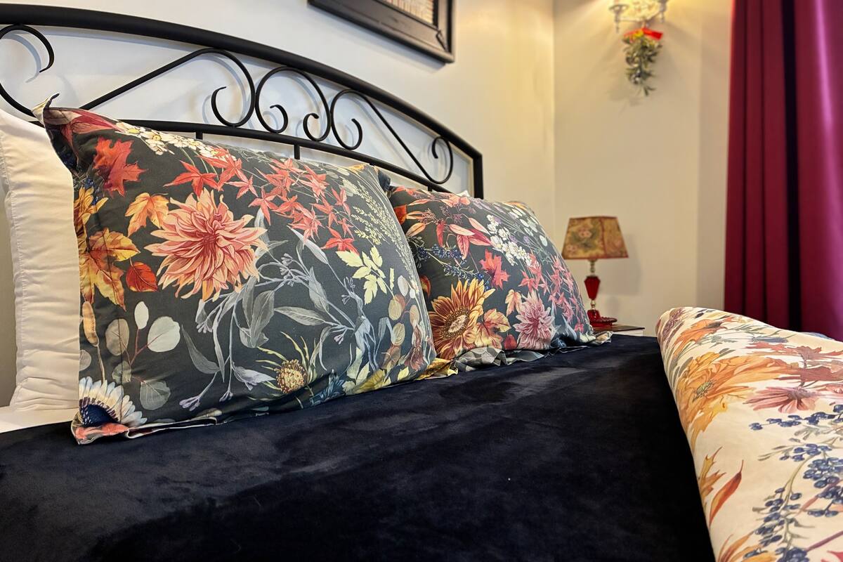 Un lit avec des oreillers à motifs floraux, une tête de lit sombre et une lampe fixée au mur.