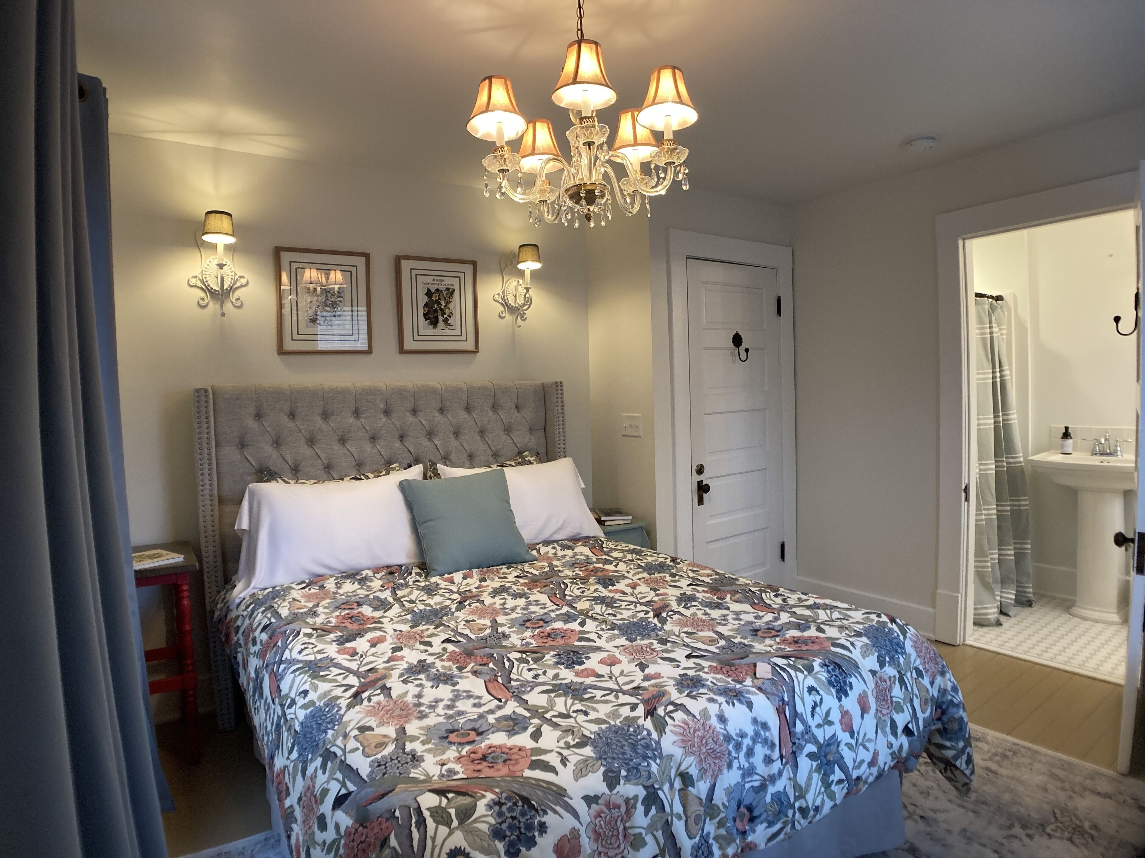 A bedroom with a large bed, a chandelier, framed pictures, and a door leading to a bathroom.