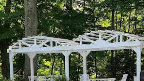 Une pergola blanche dotée de sièges extérieurs, entourée d’une végétation luxuriante et de plantes en fleurs.