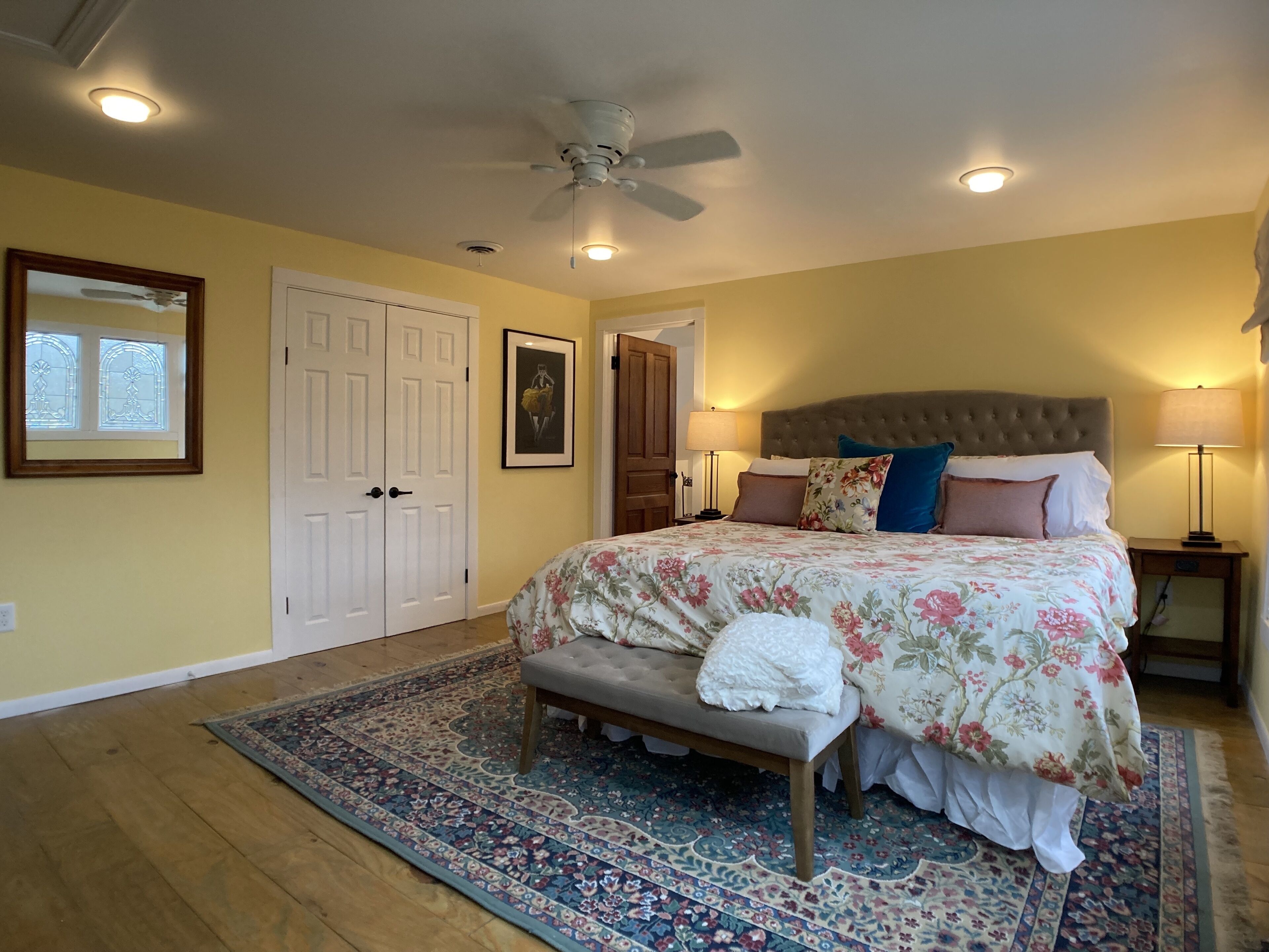A bedroom with a large bed, a bench, a ceiling fan, and a nightstand with a lamp.