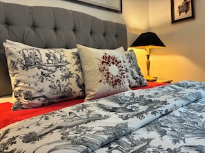 Ein Bett mit gemusterten Textilien, einem gesteppten Kopfteil und einer Nachttischlampe.