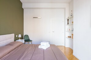 A neatly made bed with a brown comforter, a green chair, a white bedside table with a lamp, and a white door.