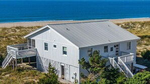 A white wooden house with a metal roof, located near a beach with a clear blue sky. - "Beach Box" BHI and Shoals Club, Pet Friendly, Ocean Views, Steps To Beach (Bald Head Island)