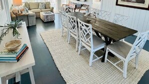 A modern living room with a wooden dining table, white chairs, a sofa, and a TV mounted on the wall. - "Beach Box" BHI and Shoals Club, Pet Friendly, Ocean Views, Steps To Beach (Bald Head Island)
