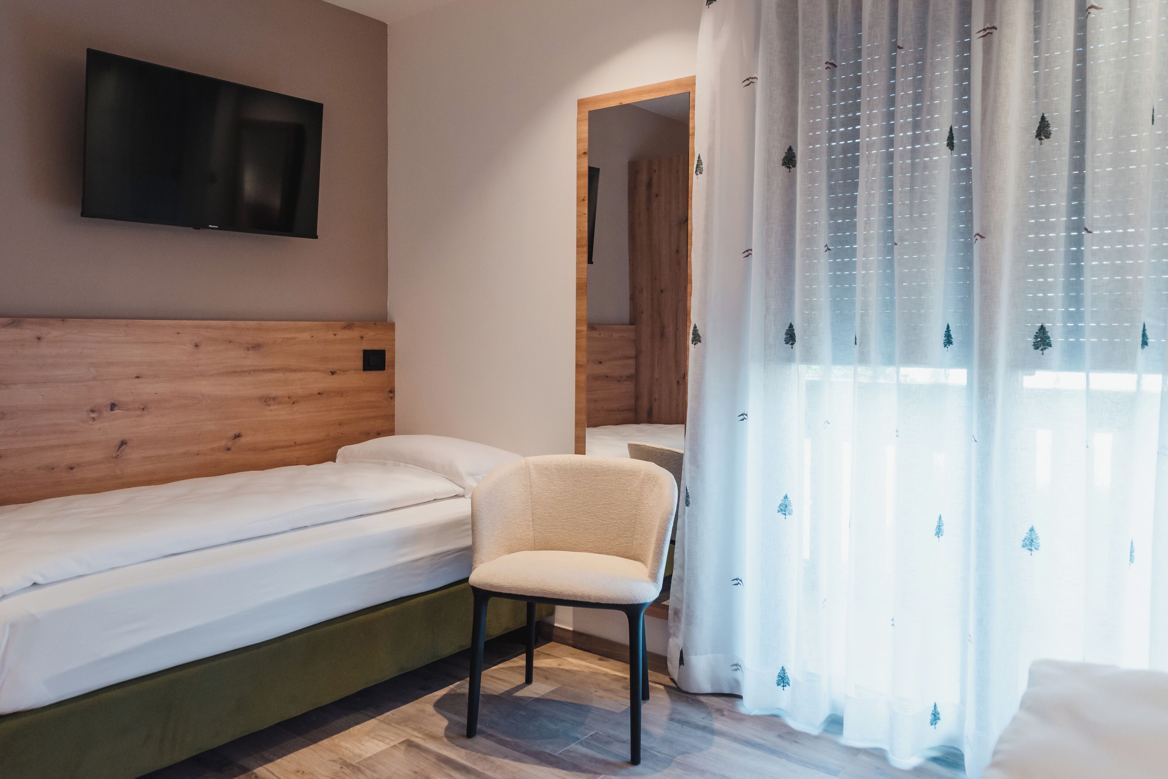 A bedroom with a bed, a chair, a TV mounted on the wall, and a window with sheer curtains.