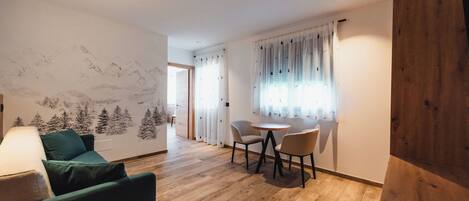 Un salón moderno con un sofá verde, suelo de madera, una mesa de comedor con sillas y un mural de montañas en la pared.
