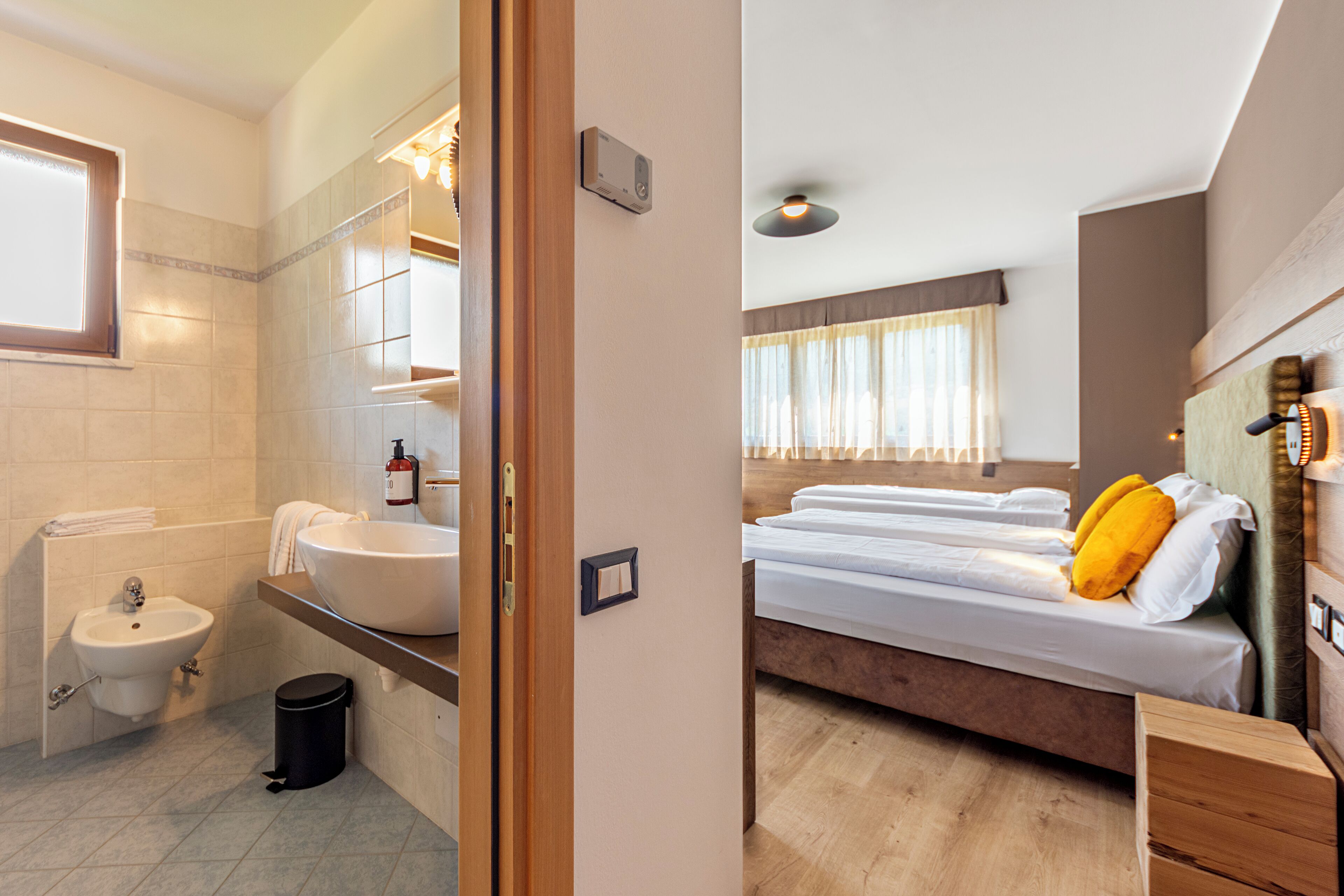 A bathroom with a sink, bidet, and a trash bin. The room has a wooden floor and a bed with white bedding and a yellow pillow.