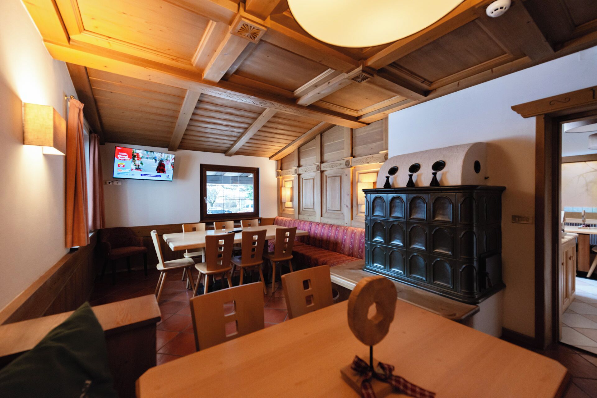 A cozy restaurant with wooden tables and benches, a television displaying a menu, and a traditional stove.