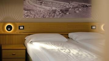 Una habitación de hotel con cabecera de madera, una cama individual con ropa blanca, una mesita de noche con cajón y una lámpara fijada a la pared.