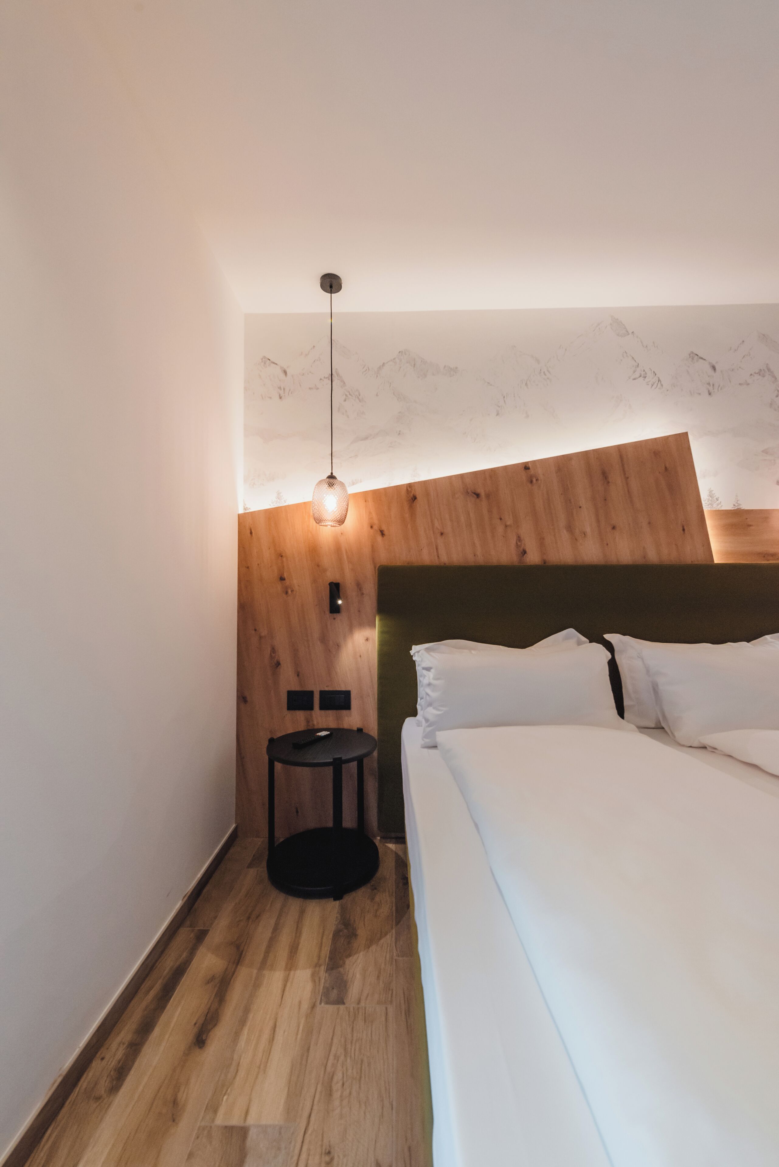 Une chambre à coucher avec une tête de lit en bois, un lit recouvert de draps blancs, une petite table ronde et un luminaire suspendu.