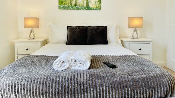 A neatly made bed with a grey textured blanket, two white towels, and a remote control on it. There are two bedside tables with lamps, a painting of a forest on the wall, and a white door in the background. - 18 Horner Street York (York)