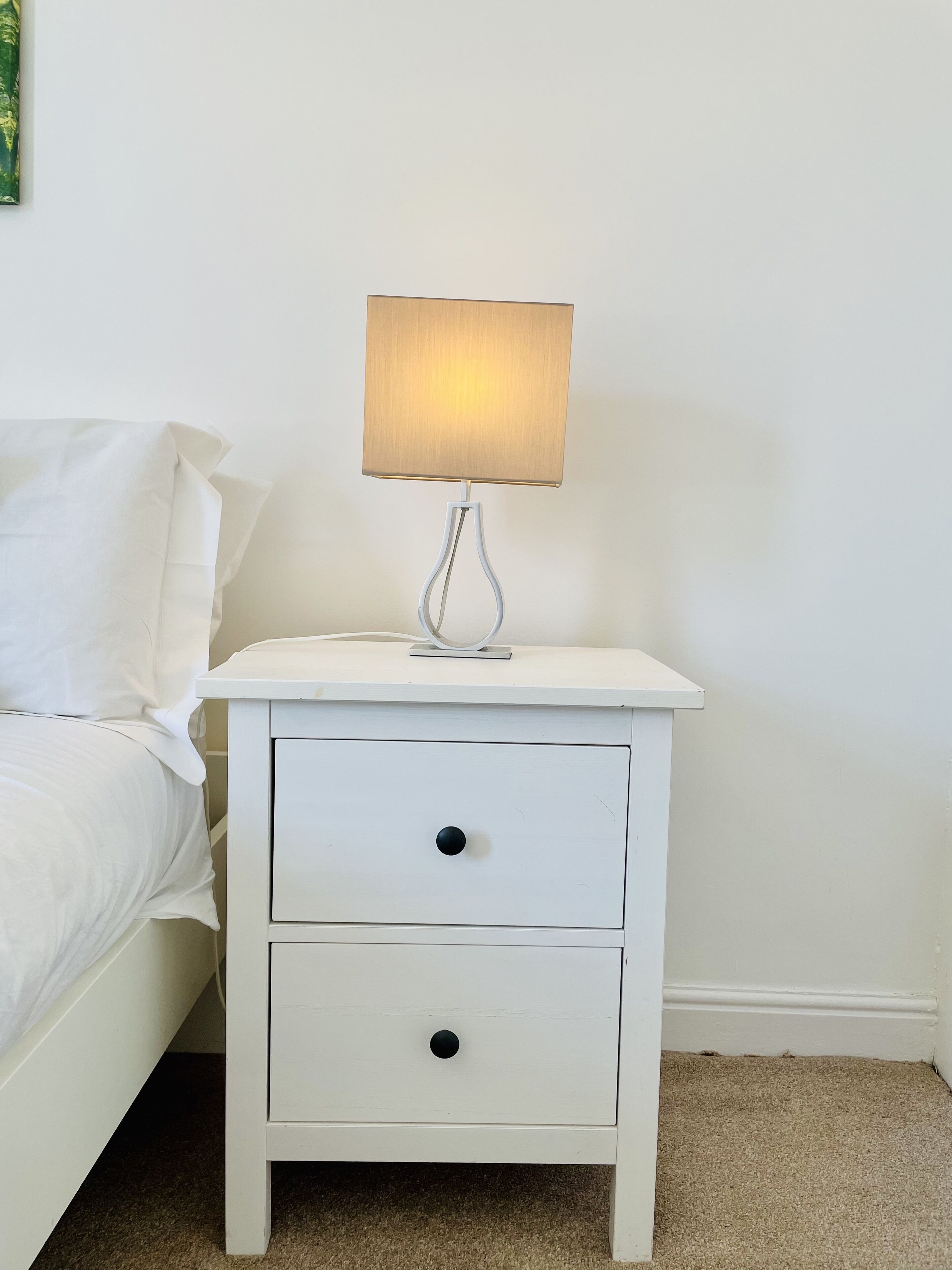 A white bedside table with a lamp, a drawer, and a bed with white bedding.