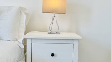 A white bedside table with a lamp, a drawer, and a bed with white bedding.
