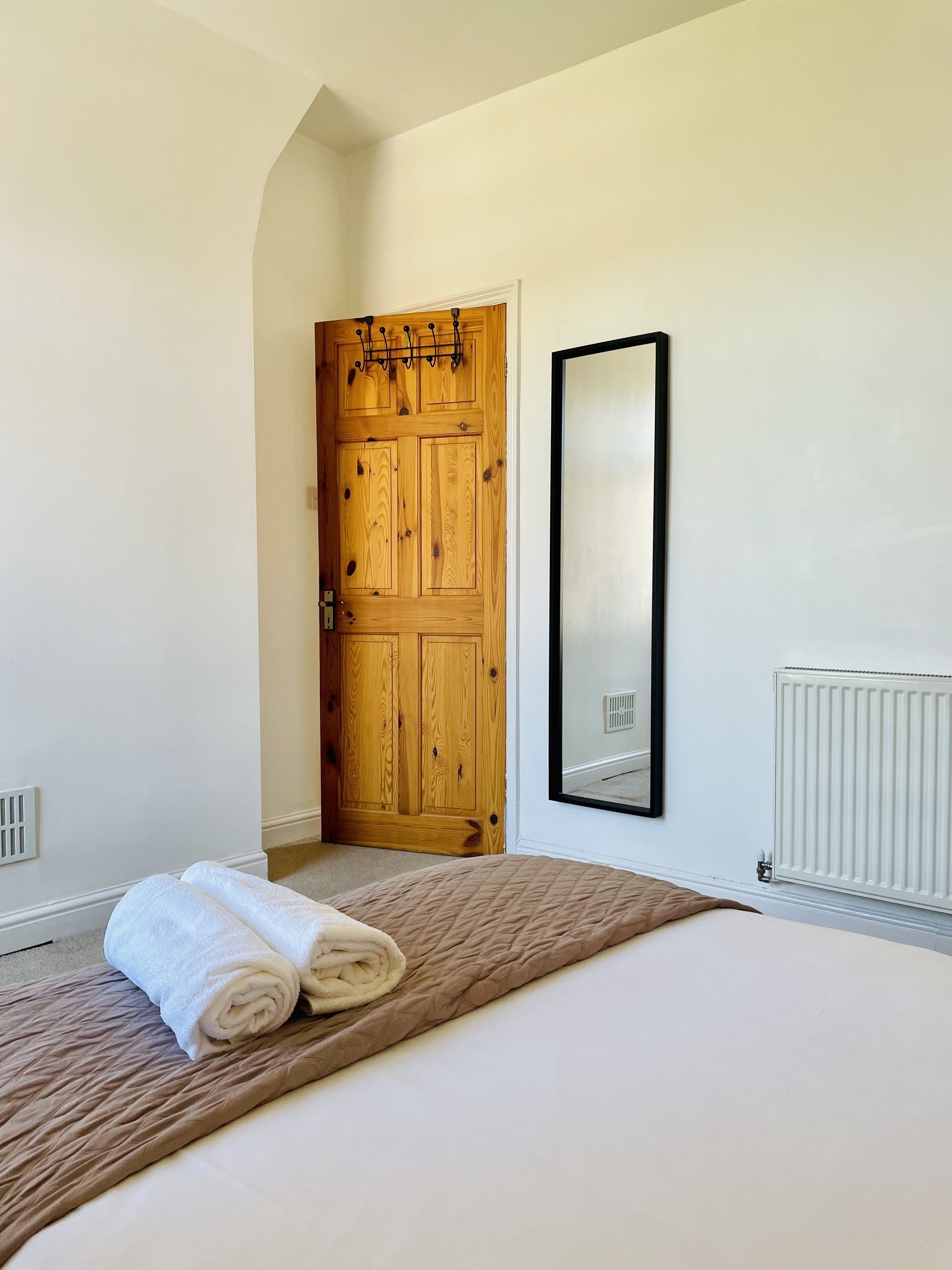 A bedroom with a wooden door, a large mirror, a radiator, and a bed with a rolled white towel.