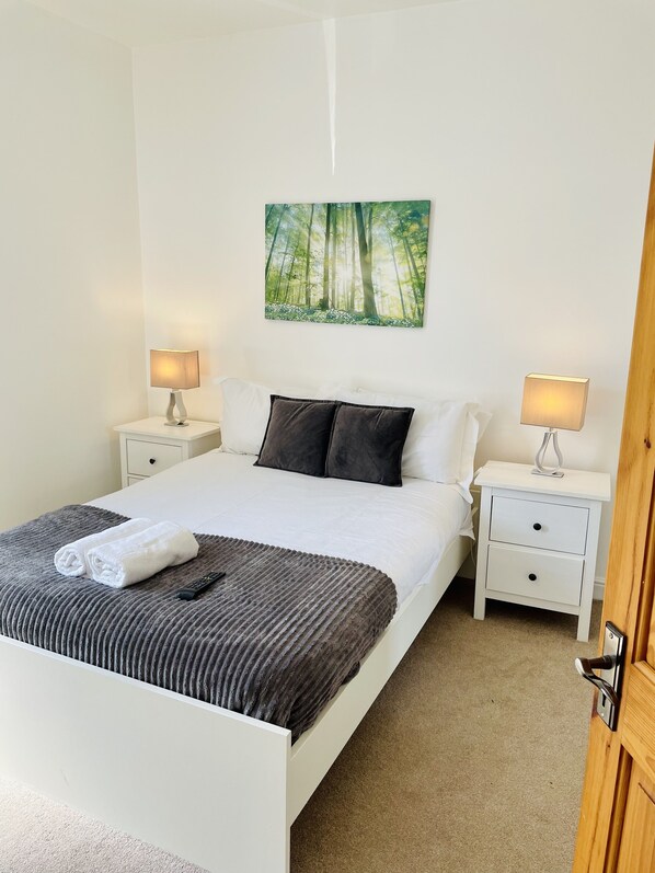 A neatly made bed with a white comforter, two pillows, and a folded towel. There are two bedside tables with lamps, a painting of a forest, and a door to the right.