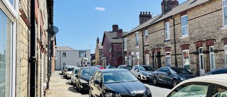 A row of parked cars along a narrow street with residential buildings on either side.