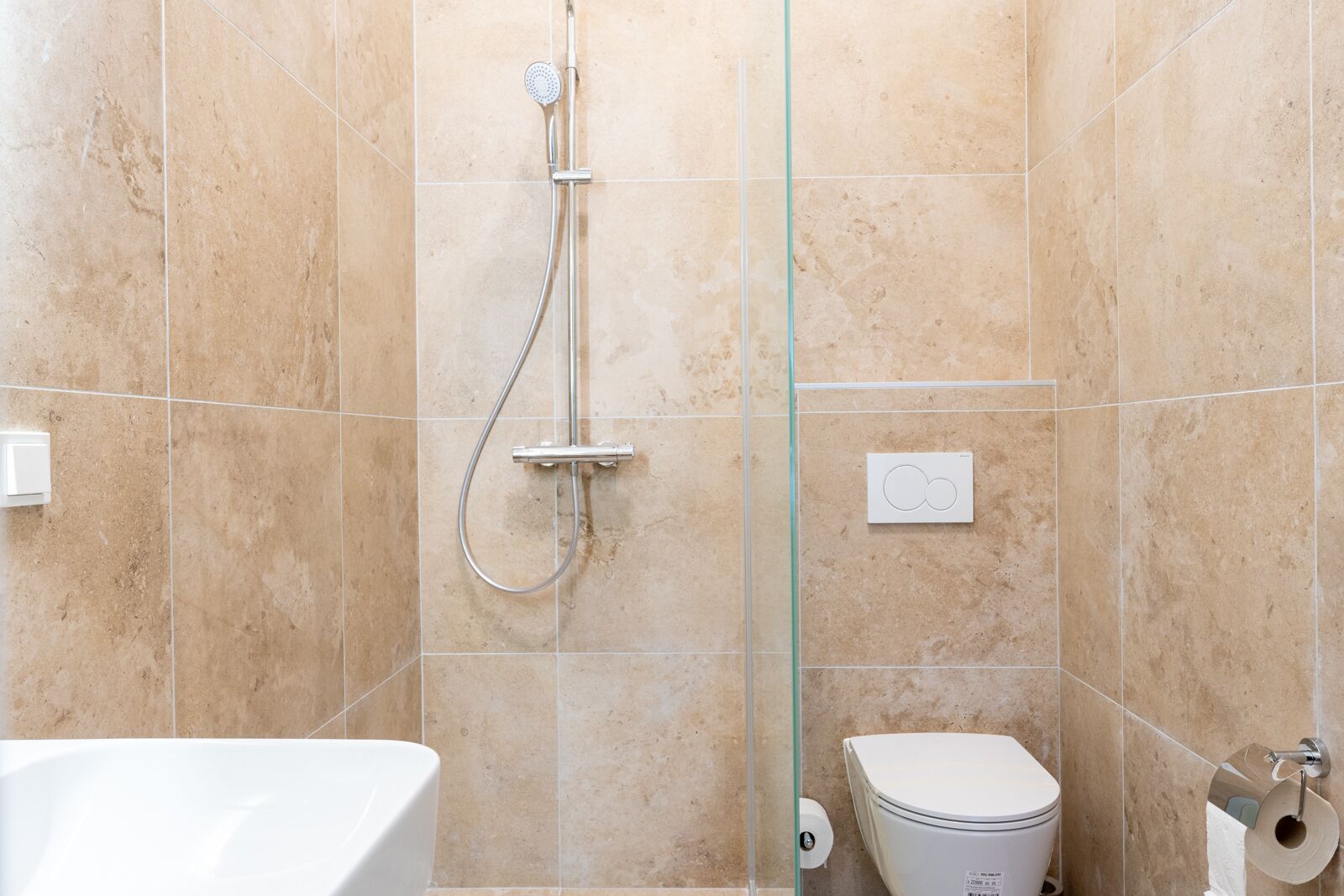 Un baño con azulejos color beige, una ducha con cabezal de ducha manual, un inodoro y una bañera blanca.