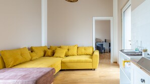 A modern living room with a yellow sectional sofa, a wooden floor, and a kitchen area with white cabinets and a countertop.