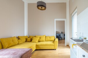 Una sala de estar moderna con un sofá seccional amarillo, piso de madera y una zona de cocina con armarios blancos y encimera.