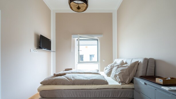 A bedroom with a bed, a TV mounted on the wall, a window, and a desk with drawers.