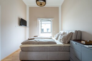 Un dormitorio con cama, televisión montada en la pared, ventana y escritorio con cajones.