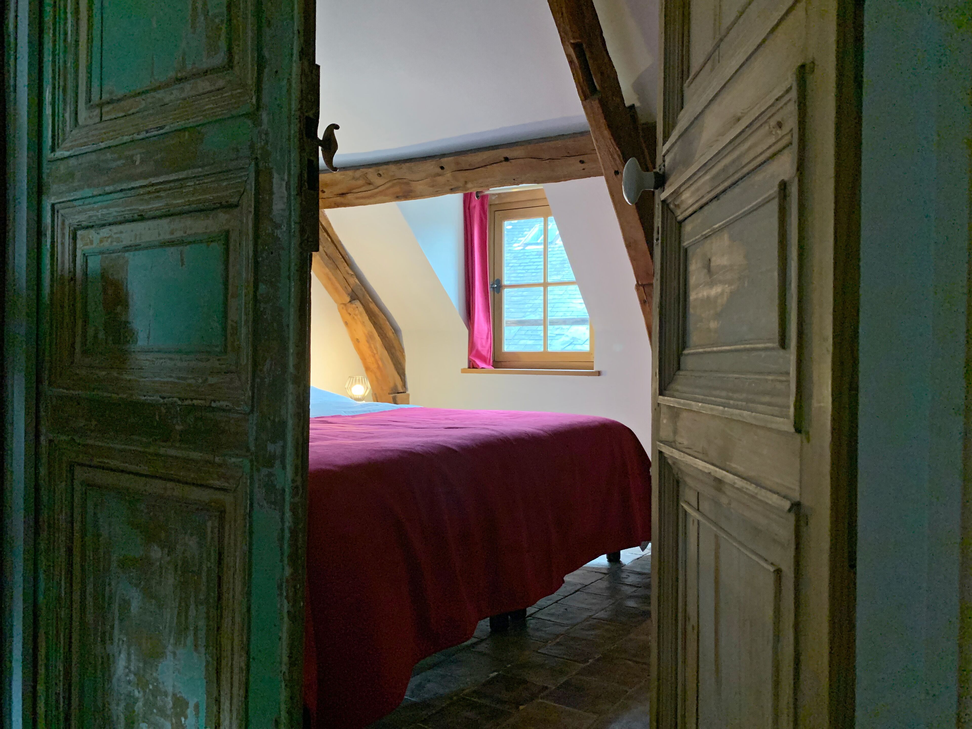 A bedroom with a wooden ceiling, a bed with a red bedspread, and a window with pink curtains.