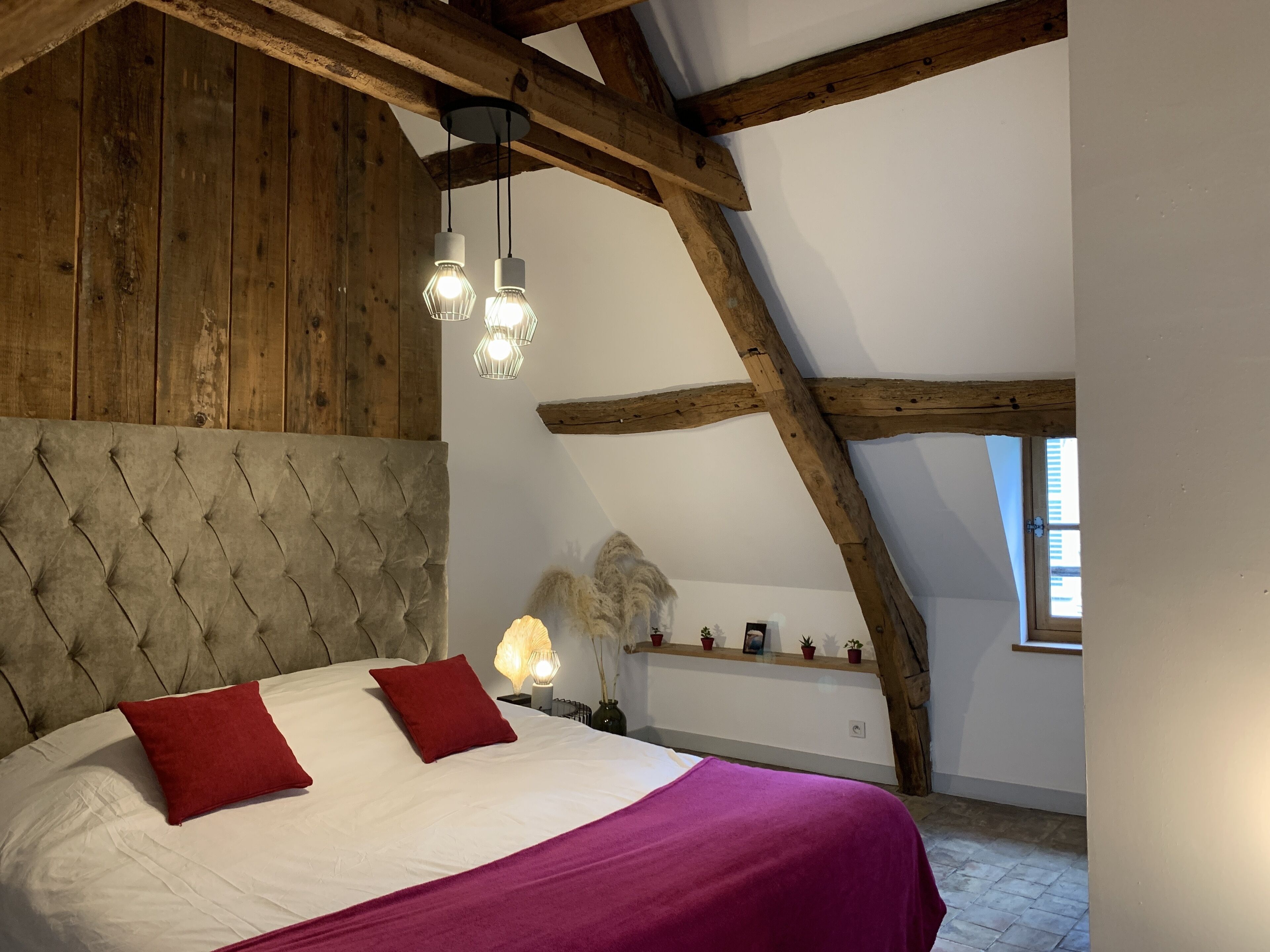A bedroom with a wooden ceiling, a tufted headboard, two red pillows, a white bedspread, and a purple blanket.