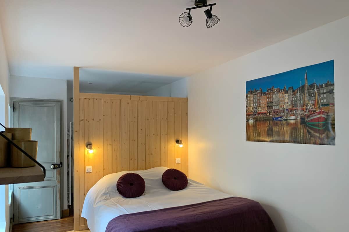 Un dormitorio con cama, cabecera de madera, dos luces fijadas a la pared, un cuadro con una vista urbana y una escalera que lleva a otra habitación.