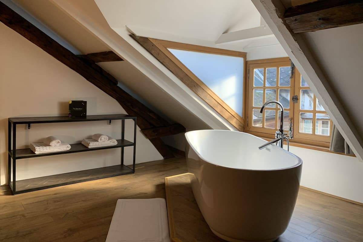 Un baño moderno con una bañera independiente, vigas de madera, un ventanal con vistas y un estante con toallas.