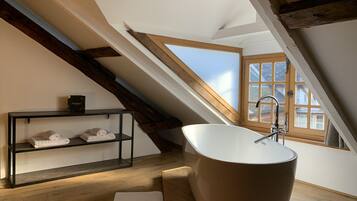 A modern bathroom with a freestanding bathtub, wooden beams, a window with a view, and a shelf with towels.