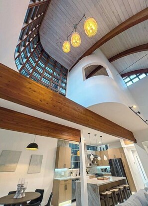 A modern interior with a high ceiling, exposed wooden beams, and a kitchen area with bar stools.